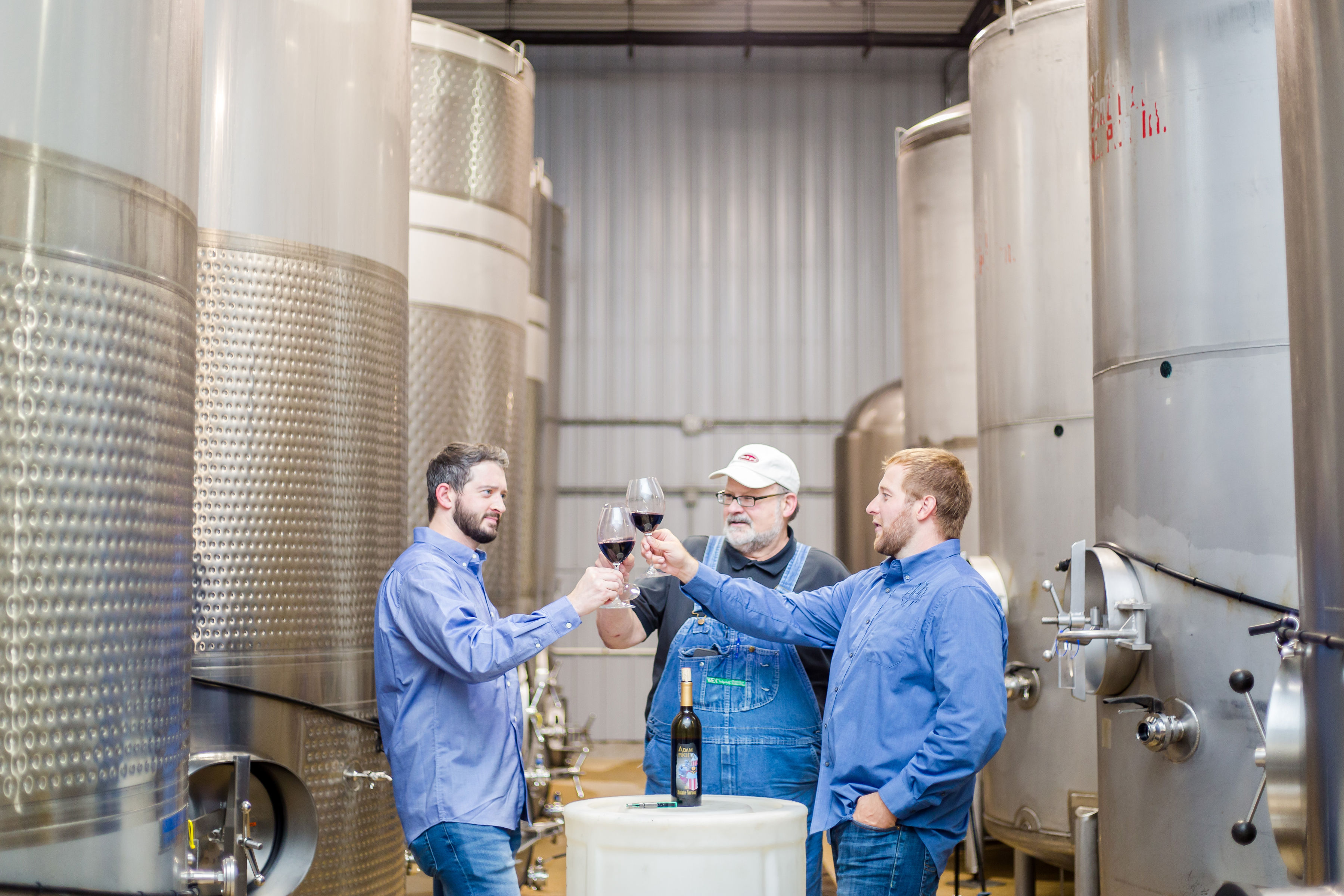 Adam Puchta Winery- Three men in blue toasting glasses of red wine. They are in a room filled with several large metal vats.