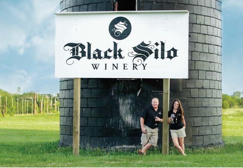 Black Silo Winery - outdoor photo, daytime, two people standing below a large sign with the winery's name. The sign is in front of a large cinder block silo.
