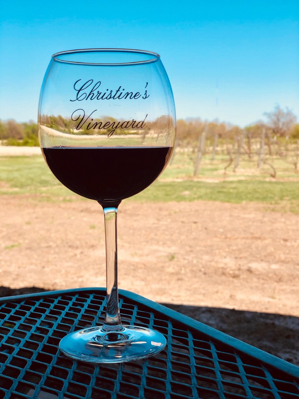 Christine's Vineyard - outdoor photo of wine glass with red wine inside it, on a patio table, with the vineyard visible in the background at daytime.