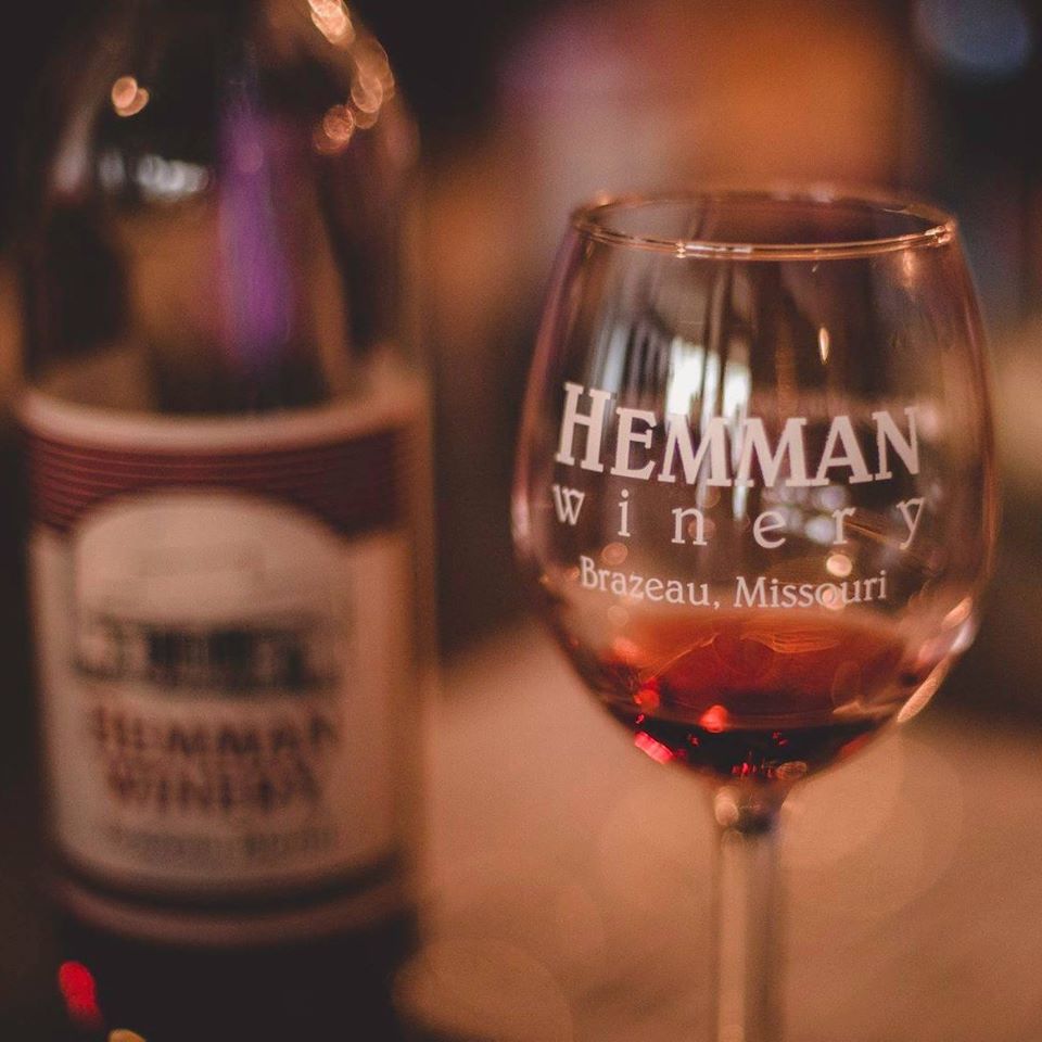 Hemman Winery- Wine glass with a small amount of red wine with a red wine bottle in the background. The wine glass is engraved with "Hemman Winery- Brazeau, Missouri".