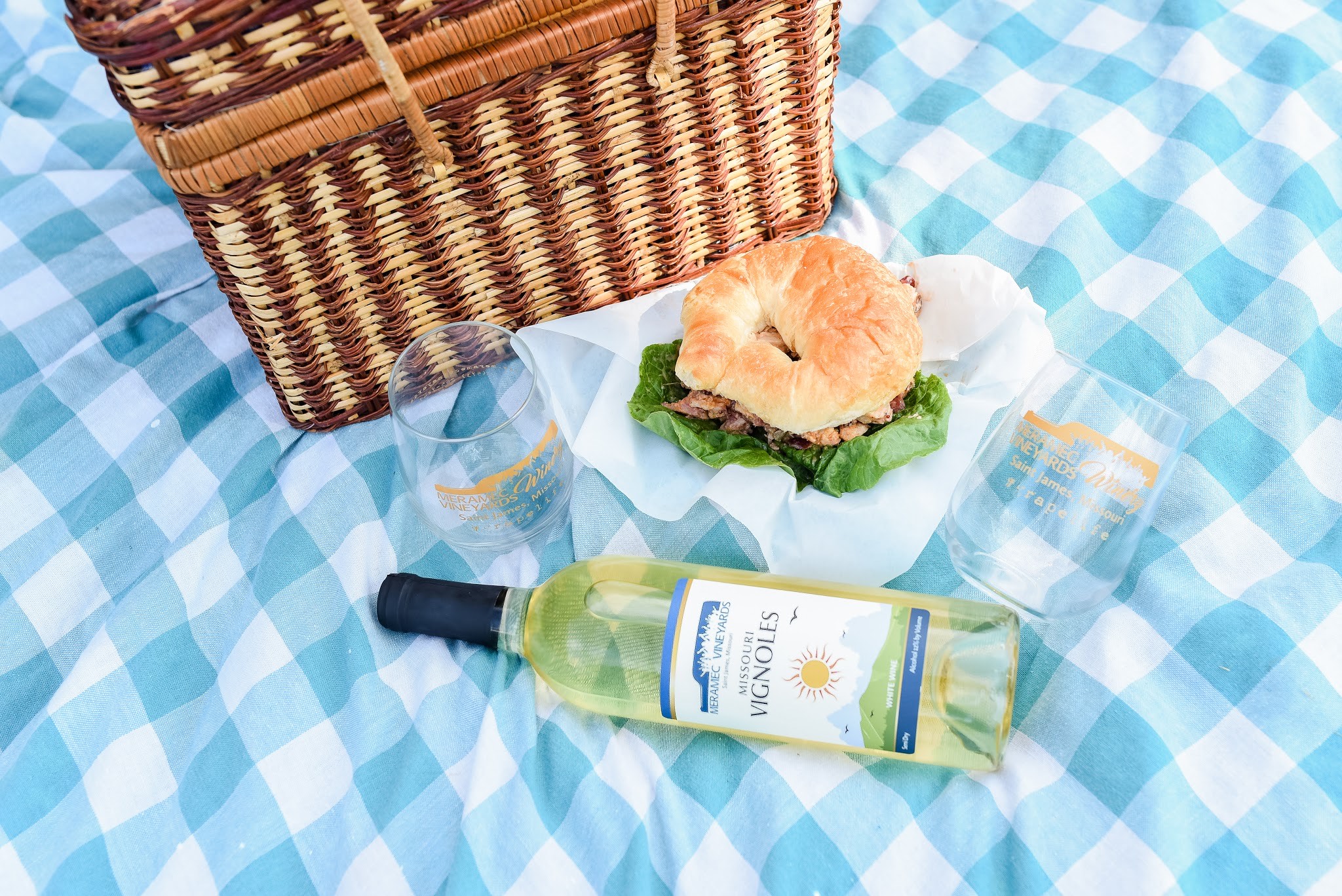 Meramec Vineyards Winery - a blue and white checkered tablecloth with a picnic basket, wine bottle, wine glass, and sandwich on it.