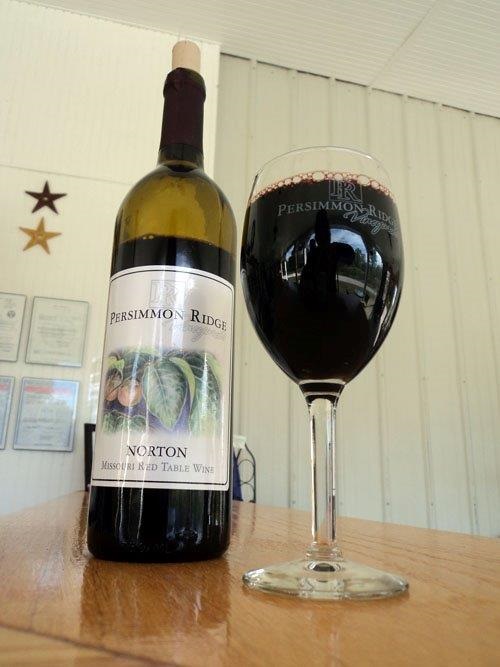 Persimmon Ridge Vineyards Winery LLC - indoor photo, a wine bottle and wine glass on a wooden table, with a white wall in the background.