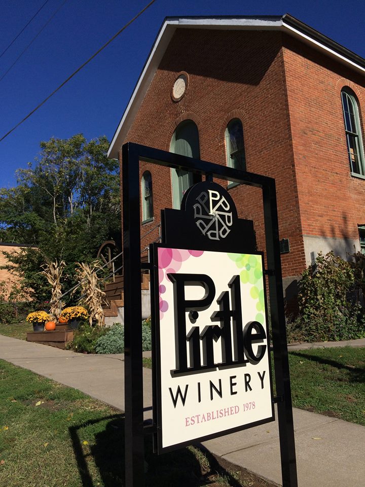 Pirtle Winery - outdoor photo, daytime, of a sign with the winery's logo. In the background is a large red brick building.