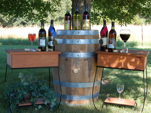 Giggling Grapes Winery - outdoor photo, daytime, of a barrel with small end tables on either side. On the barrel and tables are various wine bottles and wine glasses.