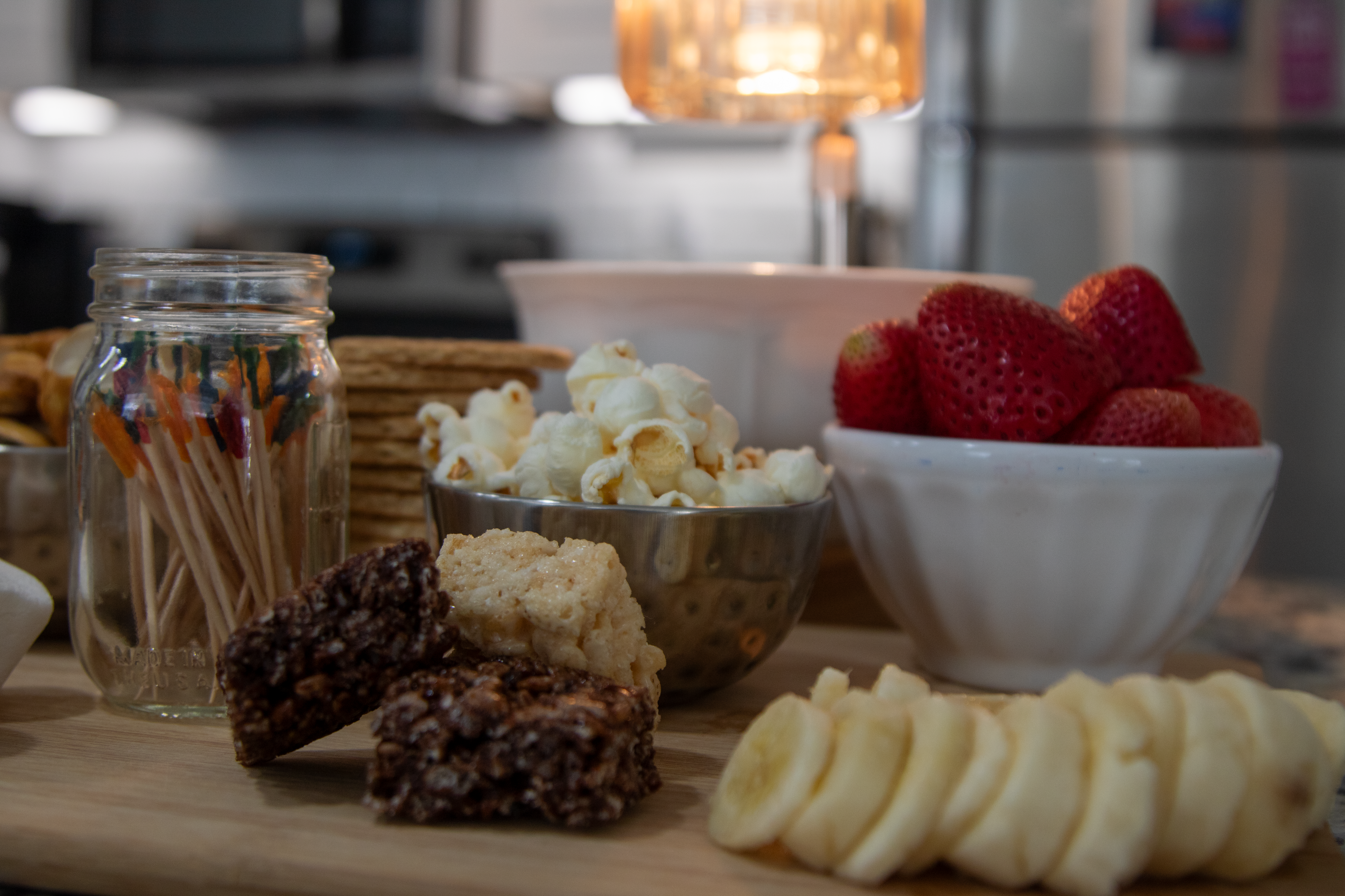 a cutting board with a variety of food on it. bananas, rice krispys, strawberries, popcorn, graham crackers and toothpicks!