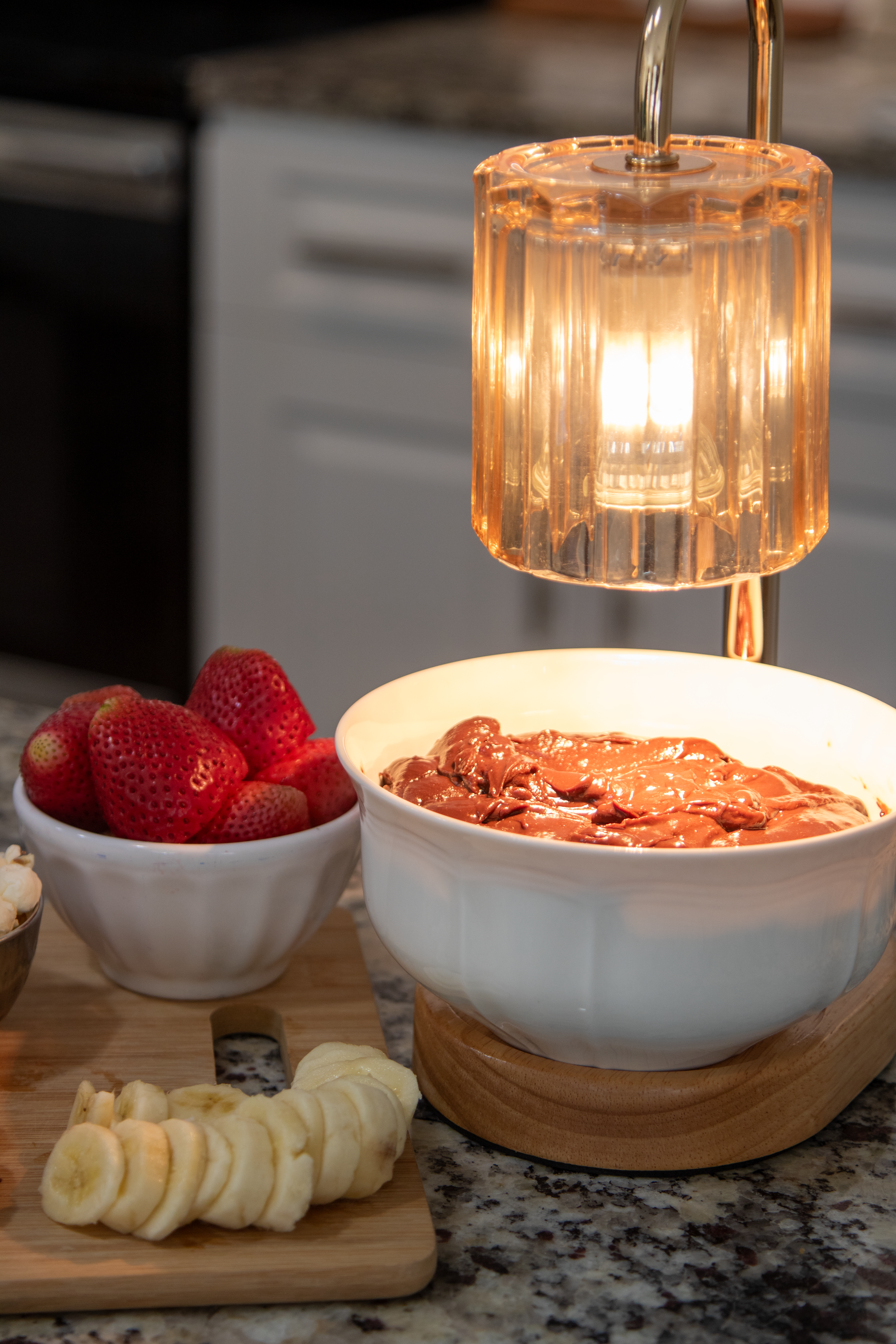 A candle warmer with a bowl of chocolate fondue sitting on it with bananas and strawberries on a cutting board next to it. 