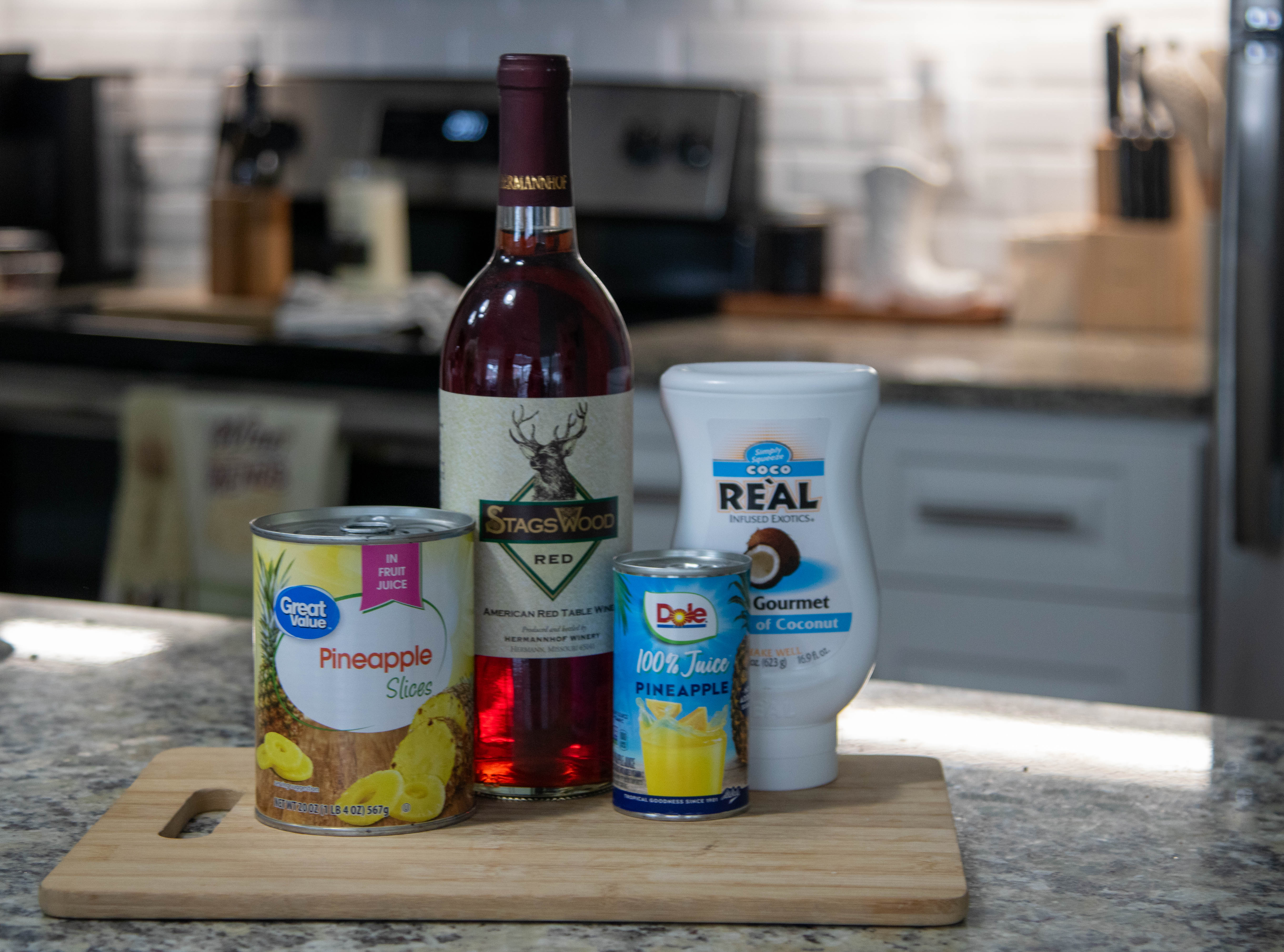 A jar of pineapple, bottle of wine, can of pineapple juice and coconut syrup sitting on a cutting board