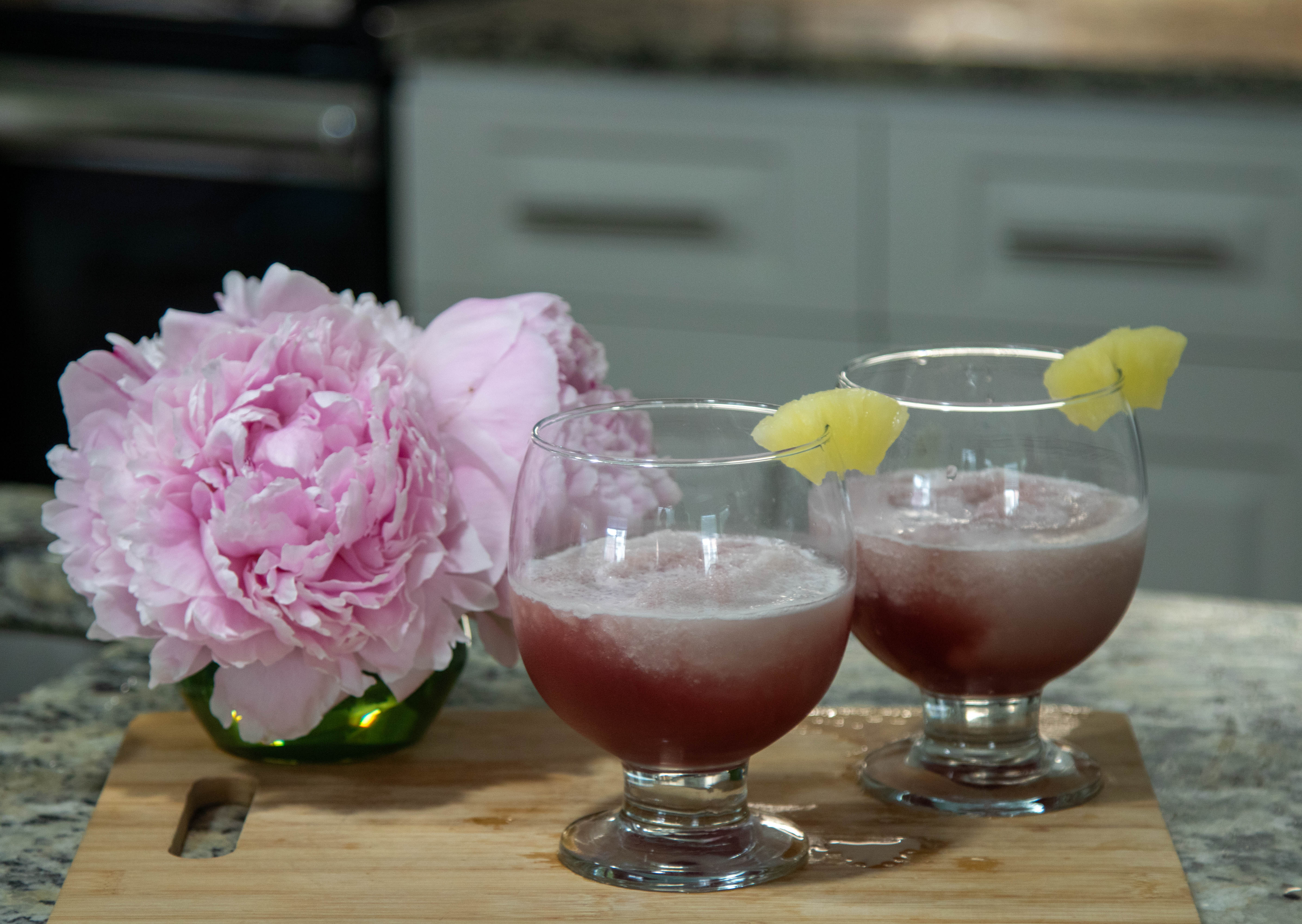 2 glasses of concord pina colada with flowers sitting on a cutting board