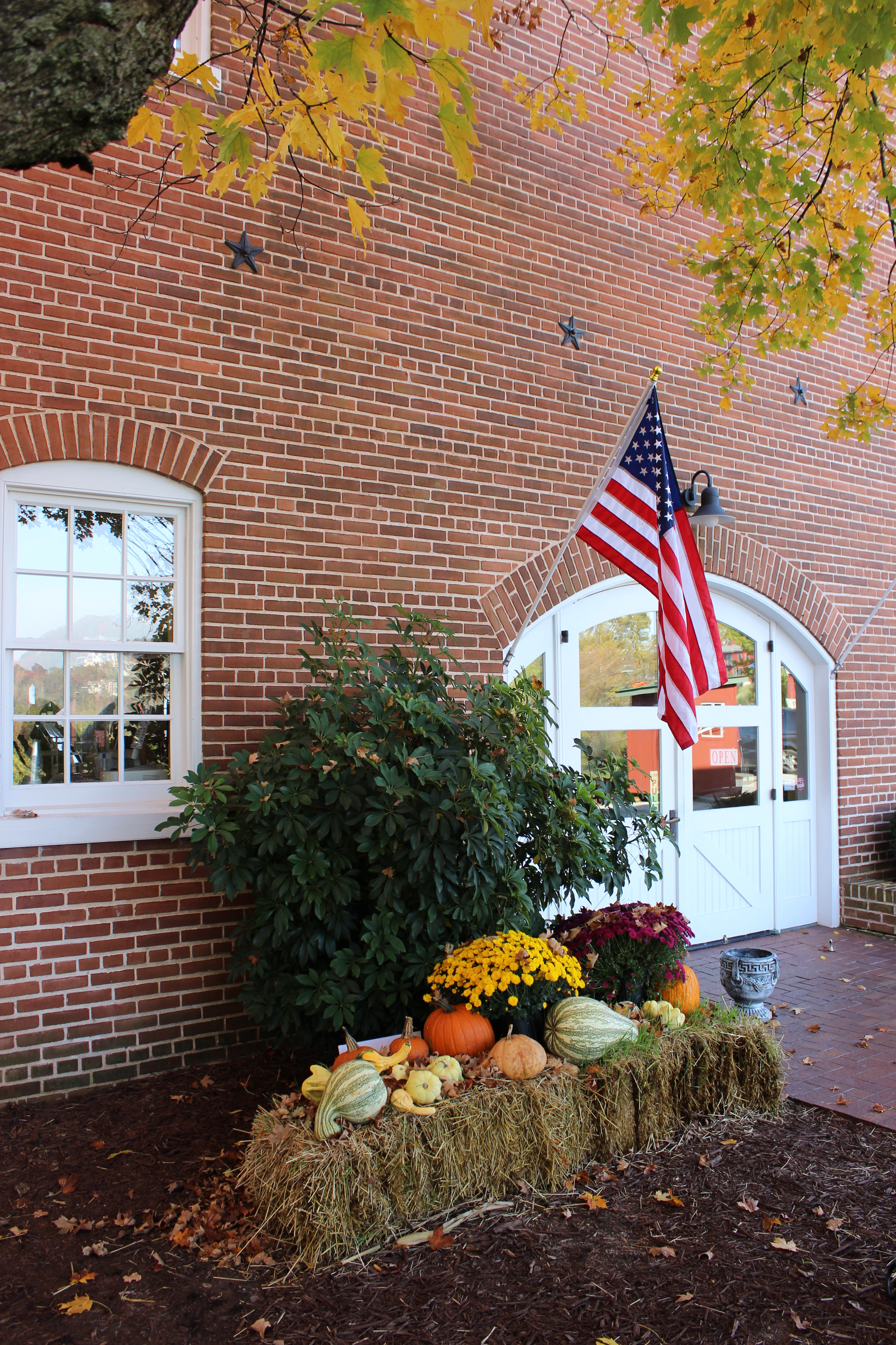 a fall setup outside a winery door that has an american flag hanging up. there are mums, pumpkins, and haybales