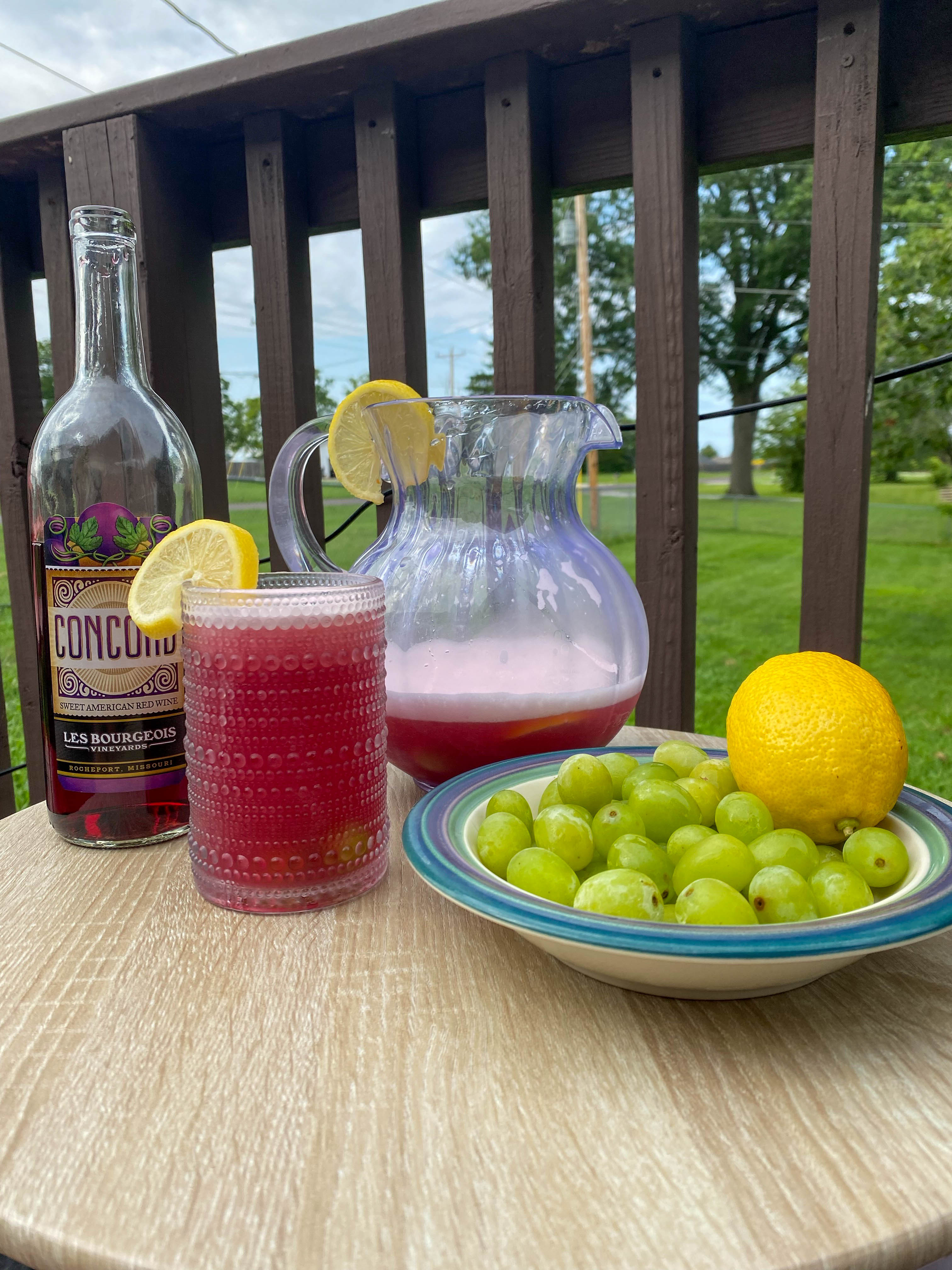 Concord Lemonade in a glass sitting on a table with a wine bottle and fruit