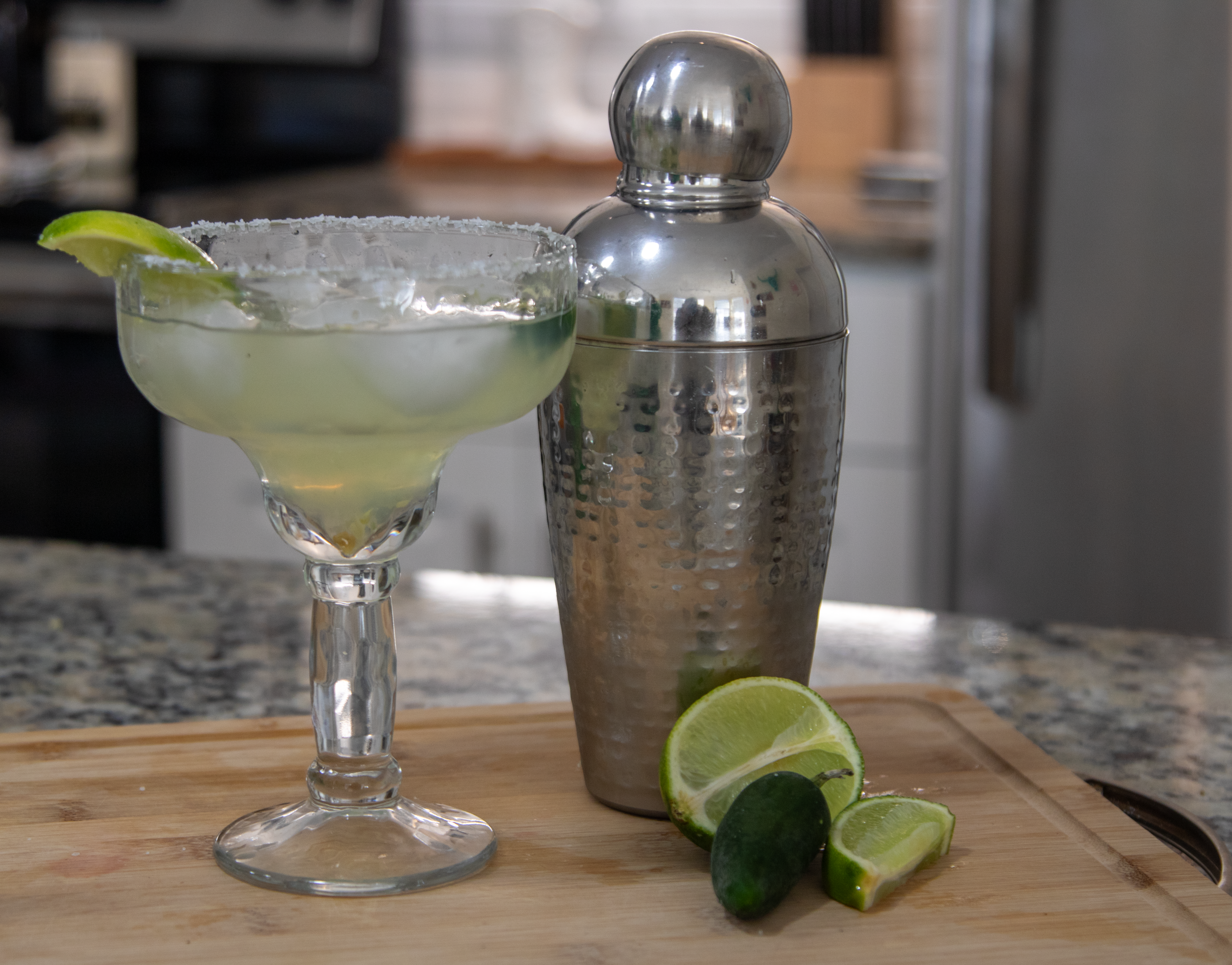 a full margarita glass sitting on a cutting board with a shaker next to it with a lime and jalapeño