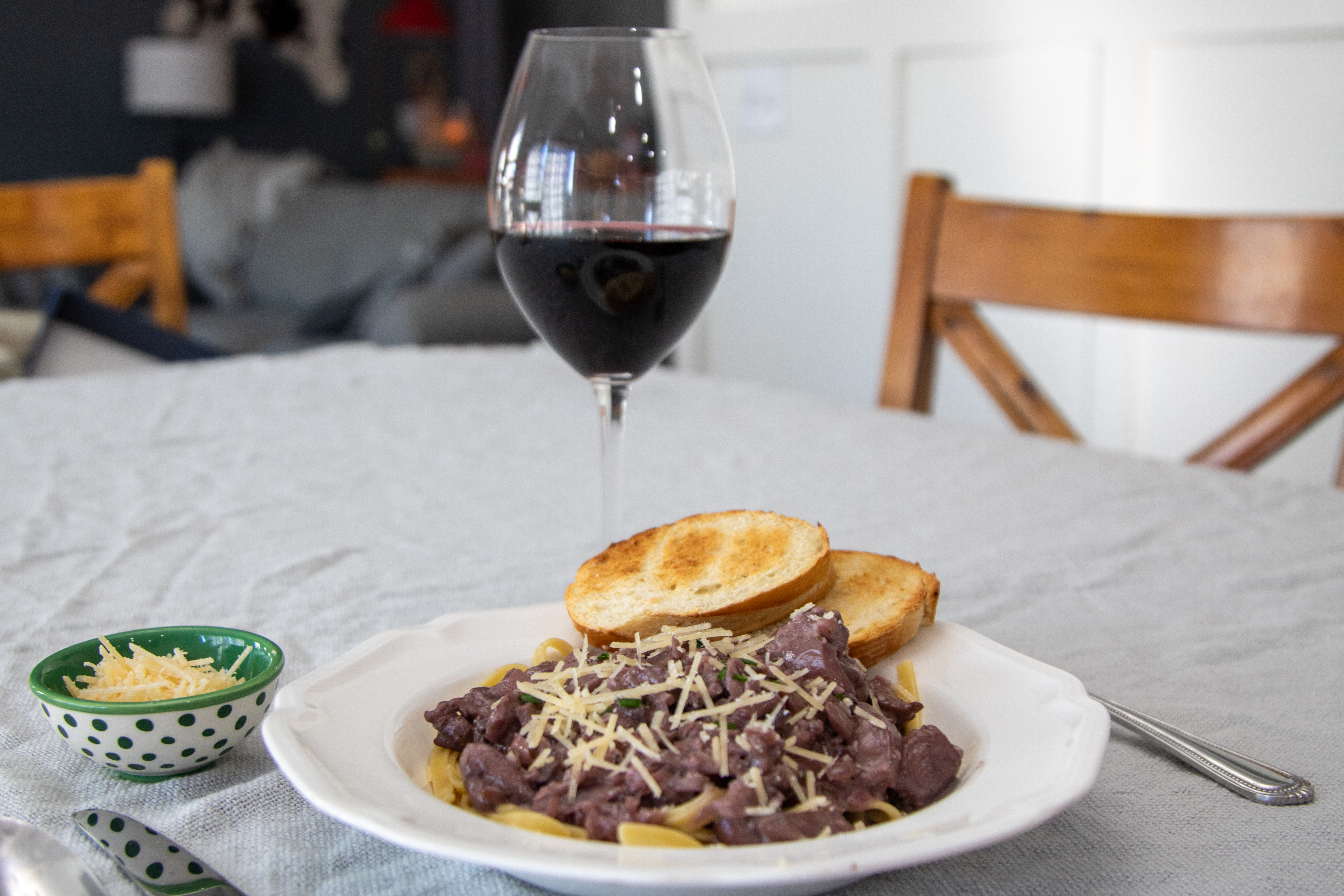 A plate of creamy chicken pasta with a slice of bread sitting on a table with a glass of red wine next to it with silverware.