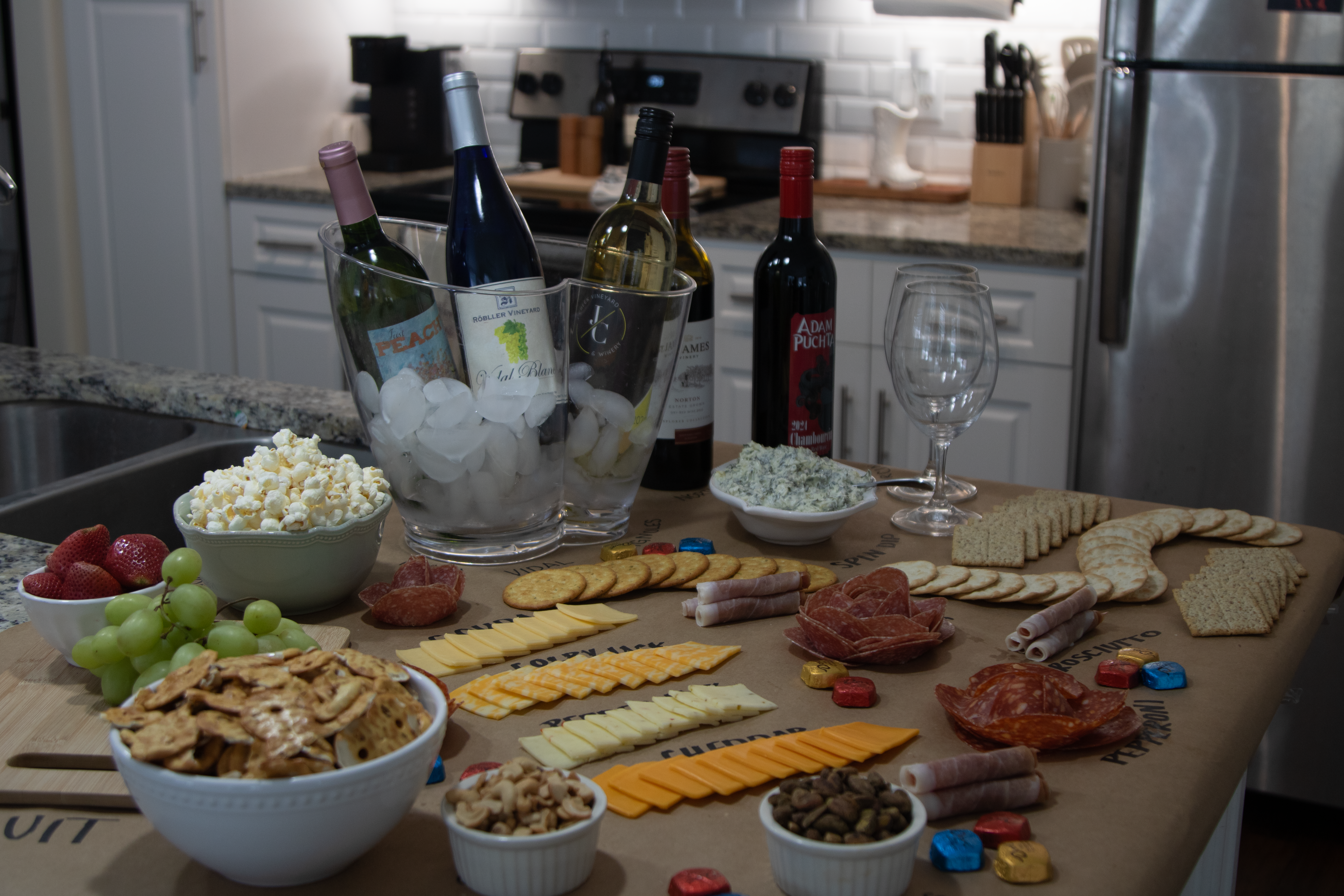 A snack board with a bunch of different snacks sitting out on a countertop with wine to pair.