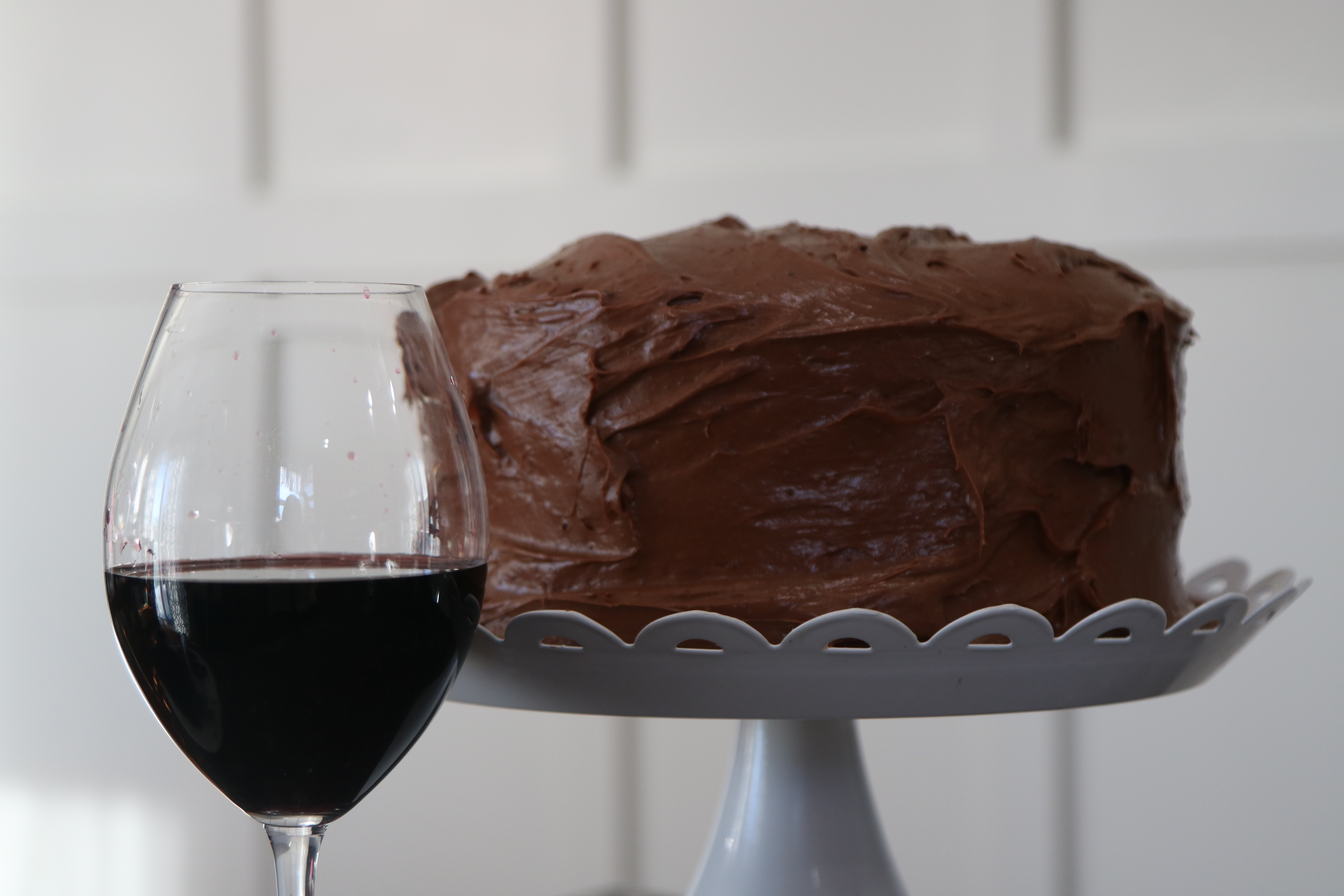 A chocolate cake sitting on a white cake stand with a glass of red wine in front of it!