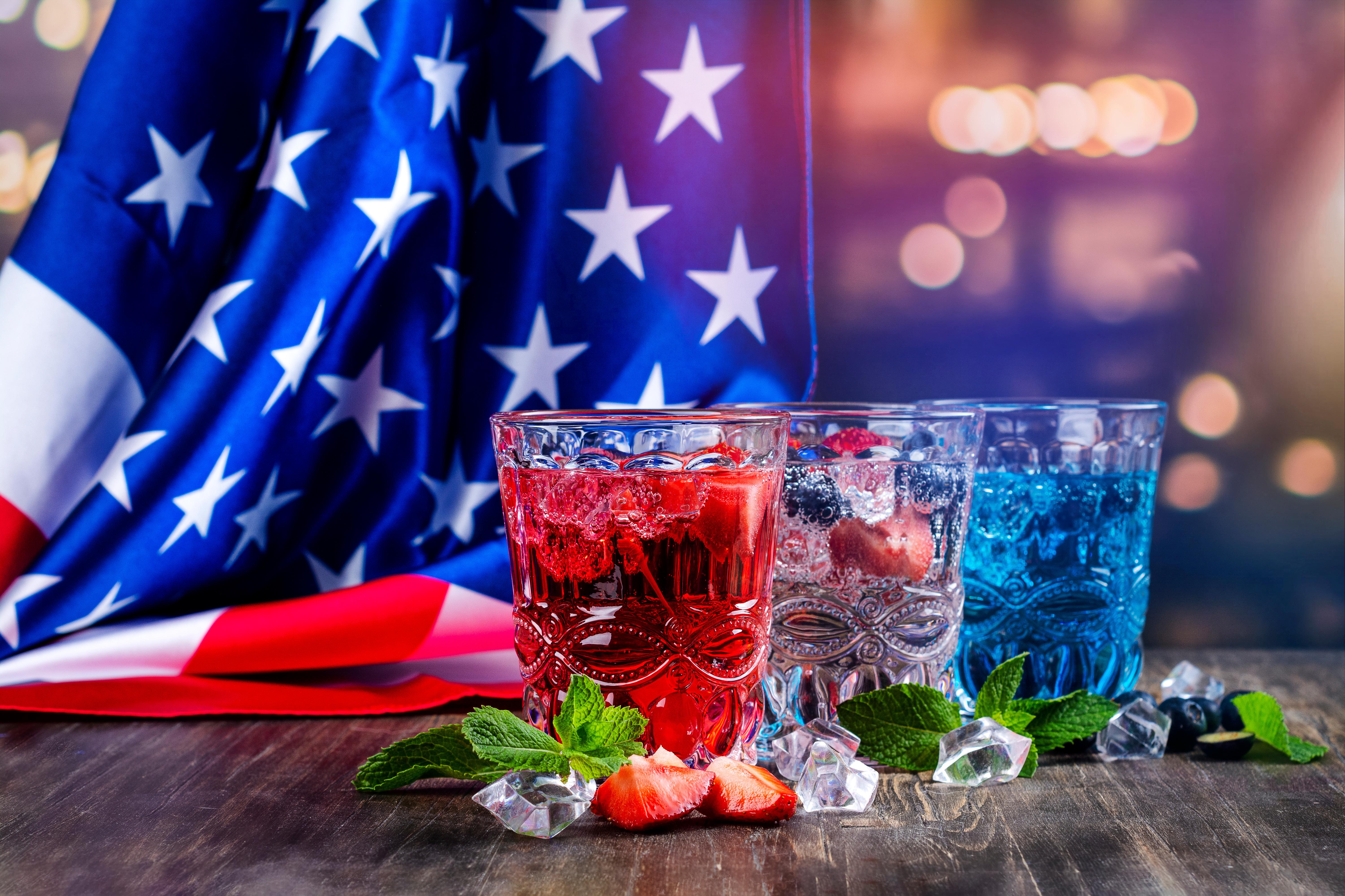 American flag hanging in the background with 3 glasses sitting in the front with red white and blue wine in them 