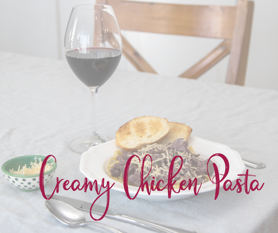 A plate of creamy chicken pasta with a slice of bread sitting on a table with a glass of red wine next to it with silverware with text over laid reading creamy chicken pasta.