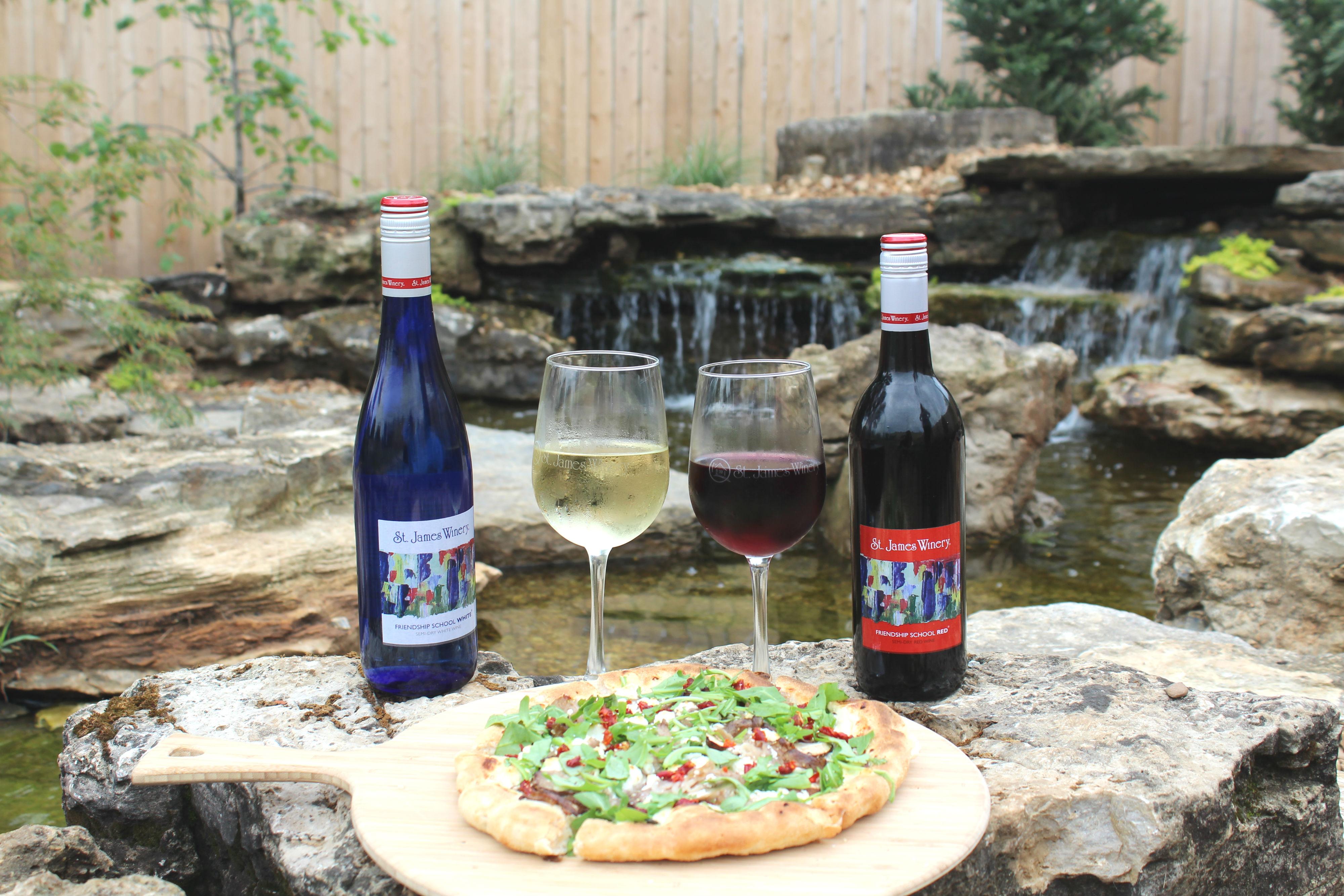 St. James Winery - Branson - outdoor photo, daytime, a plate with a pizza is on a rock with several bottles of wine in front of a small waterfall.