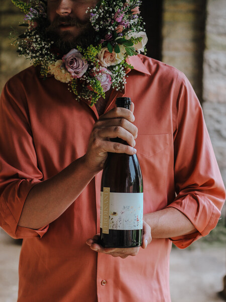 Eagles’ Landing- A man holding a bottle of red wine. He is wearing a salmon shirt with several roses and baby's breath flowers in his beard.